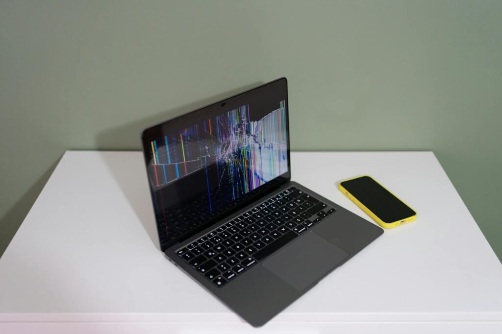 A damaged laptop screen with colorful lines and a yellow smartphone on a white desk.